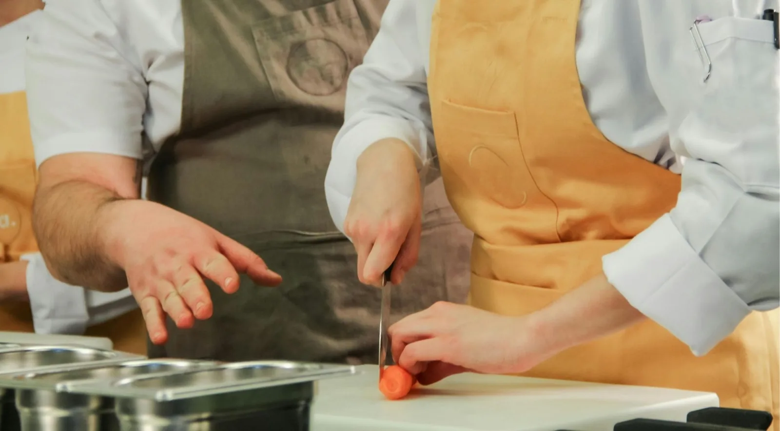 A chef guiding a guest's knife work at a stainless steel prep counter