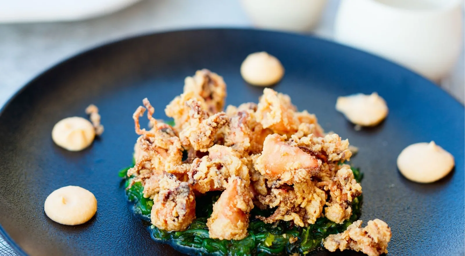 Crisp fried calamari served over wilted greens with dots of pale sauce on a dark plate