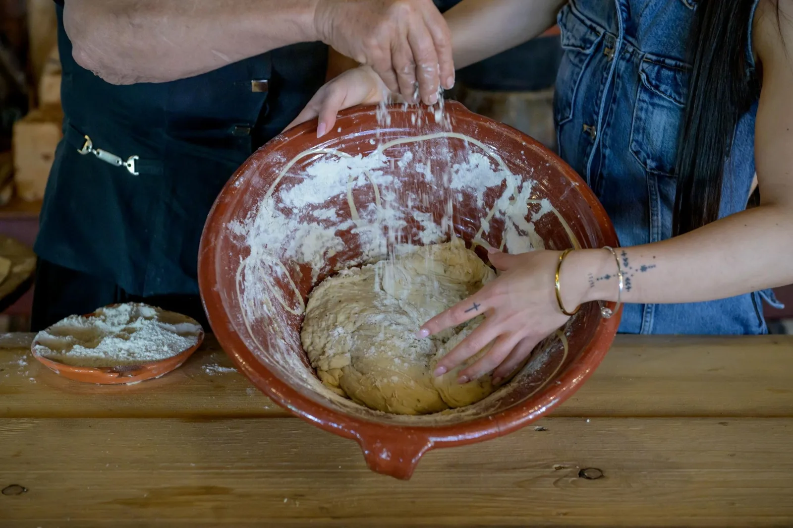 Two pairs of floured hands kneading bread dough together in a large terracotta bowl