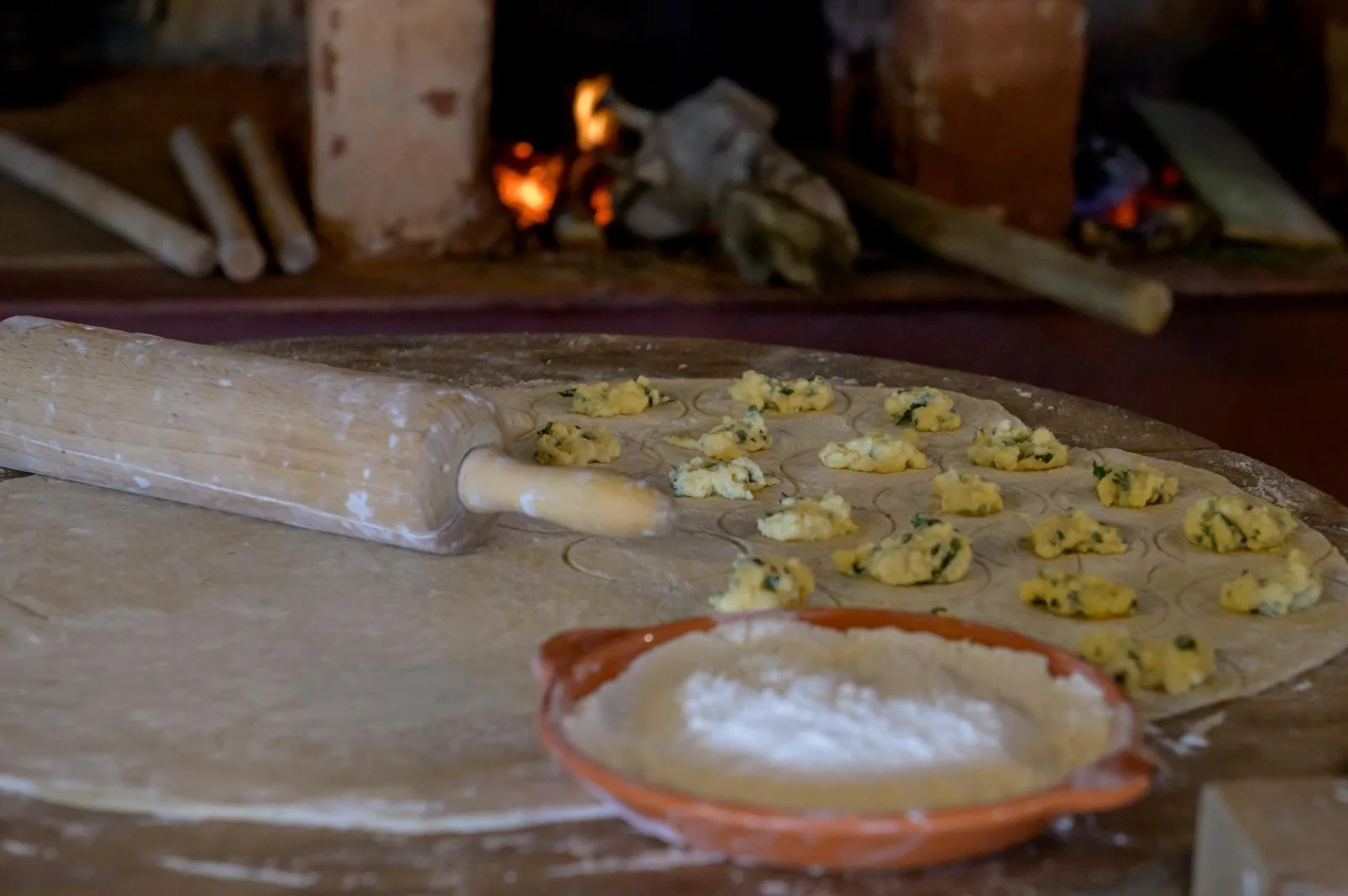 A lit wood-fired oven behind a floured board of filled pastries and a rolling pin