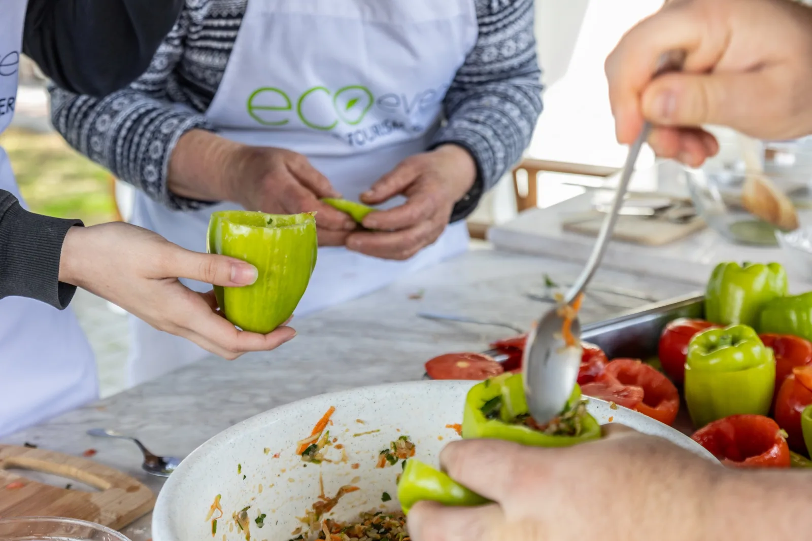 Hands spooning a rice and herb filling into hollowed green peppers beside a tray of tomatoes