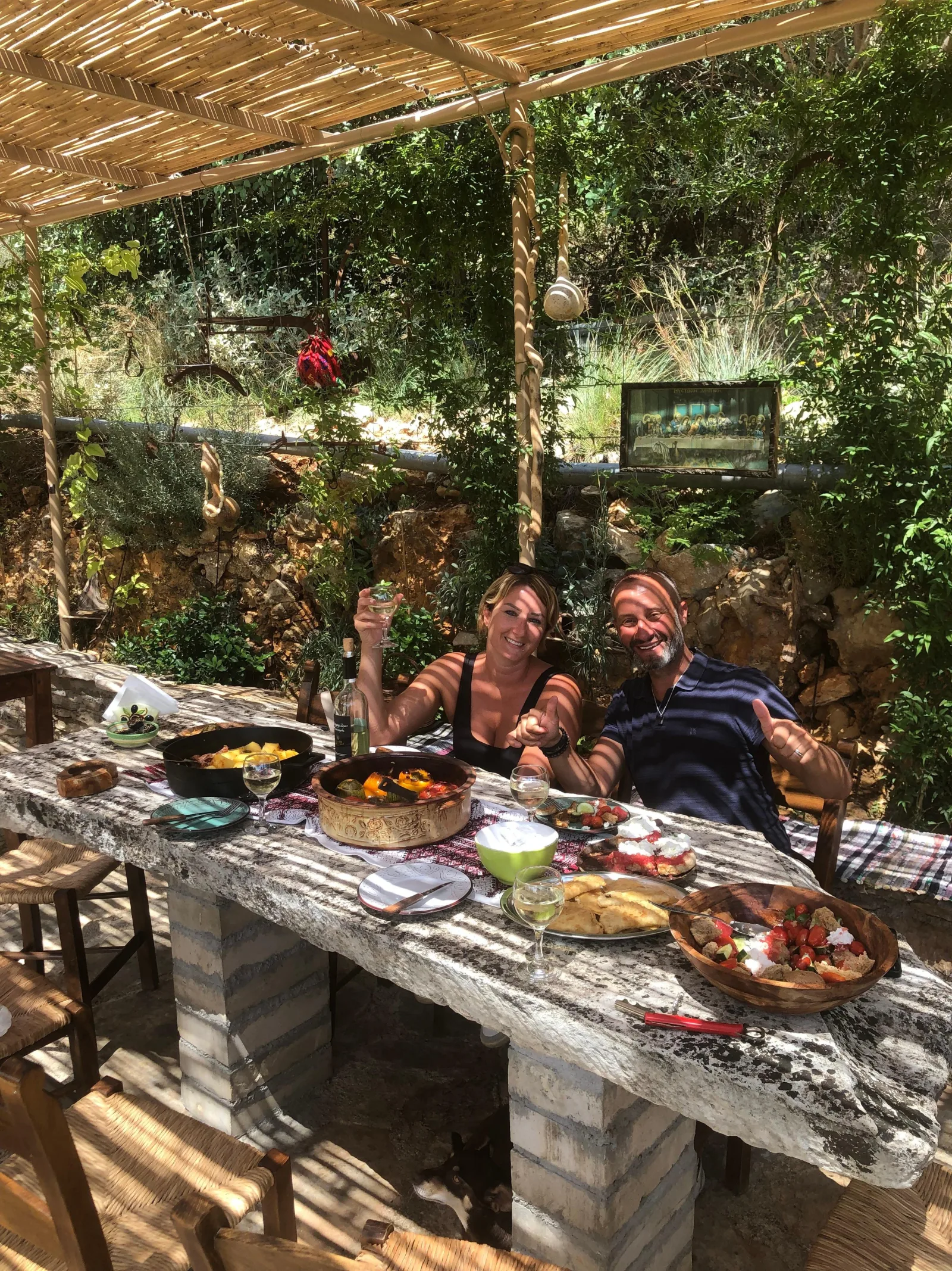 Guests eating together at a long stone table under a vine-covered pergola on a Cretan farm