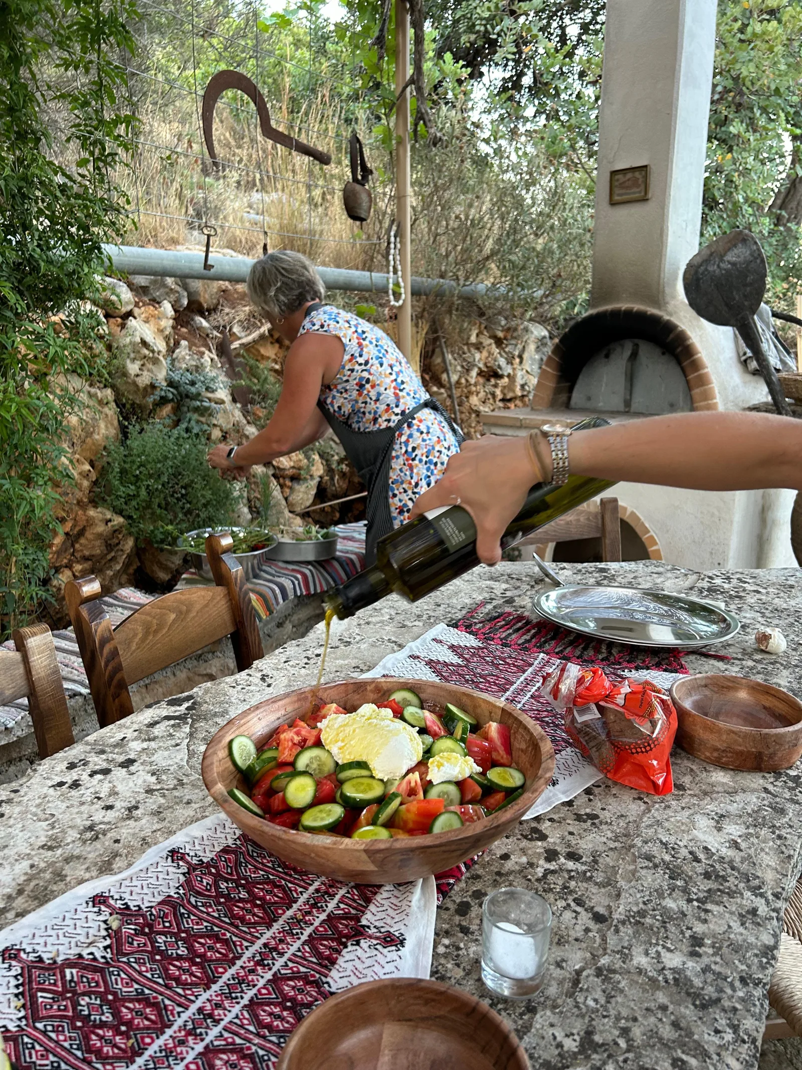 Wine being poured at an outdoor table set with a woven runner and a bowl of Cretan salad, a stone wood oven behind