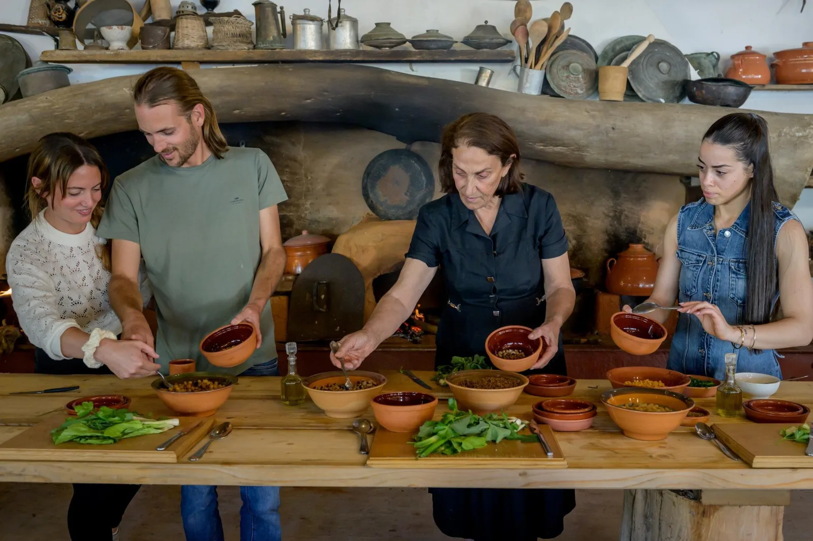 Four guests working with clay vessels and bowls of legumes and greens at a long wooden bench