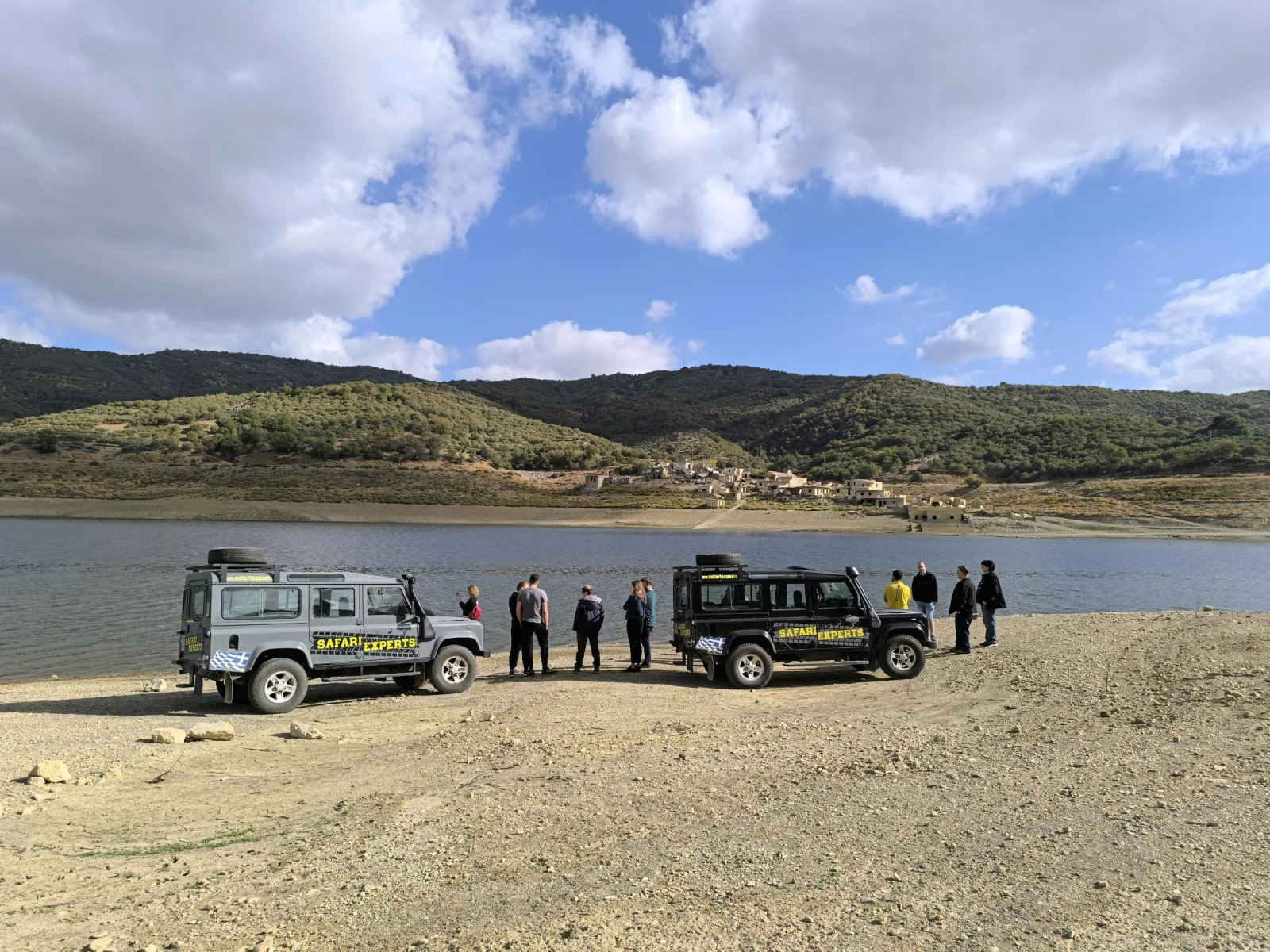 Two Land Rover 4x4s parked at the shore of a reservoir with the group standing beside them