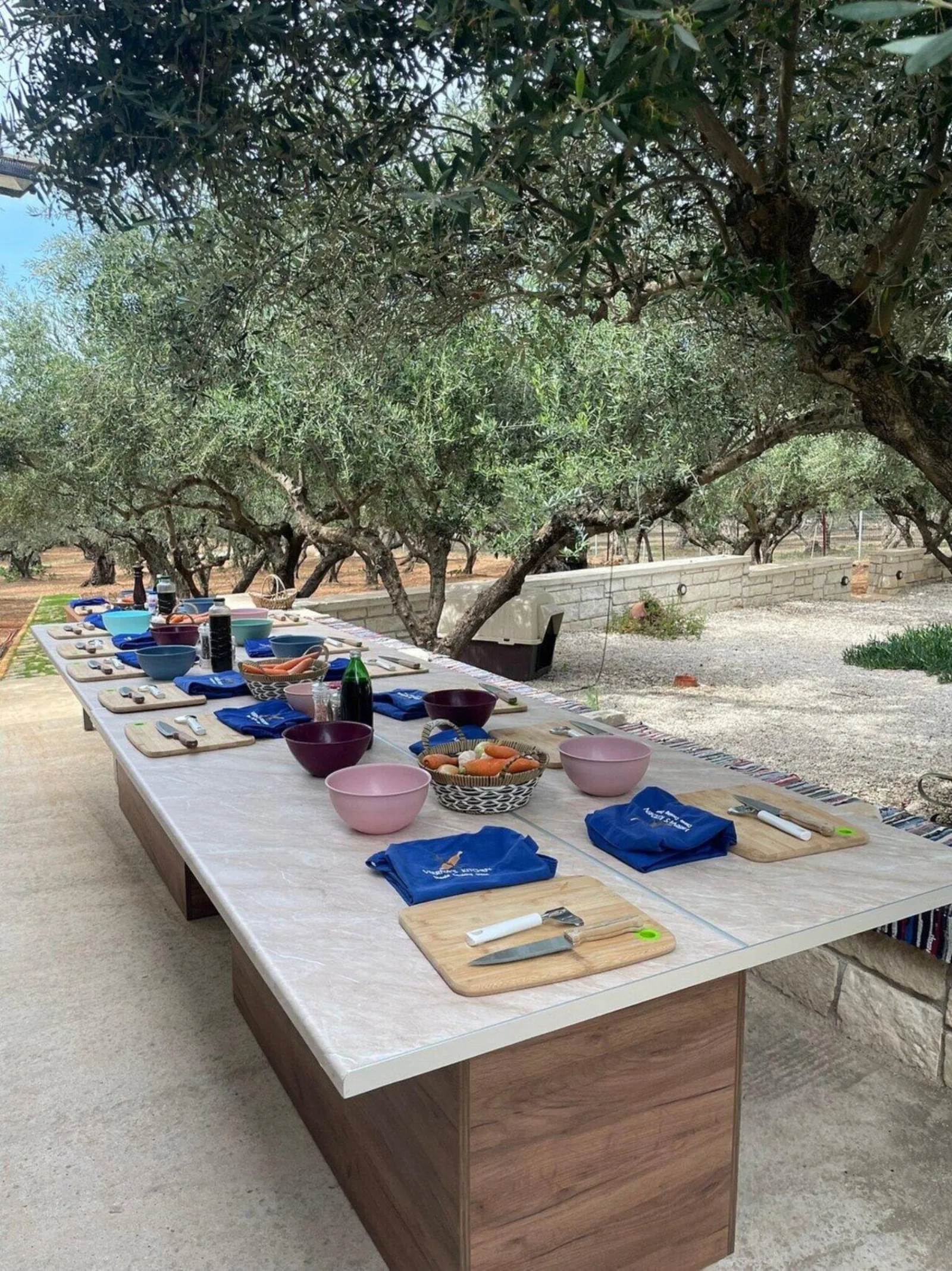 A long marble prep table set with placemats and mixing bowls under olive trees in the garden
