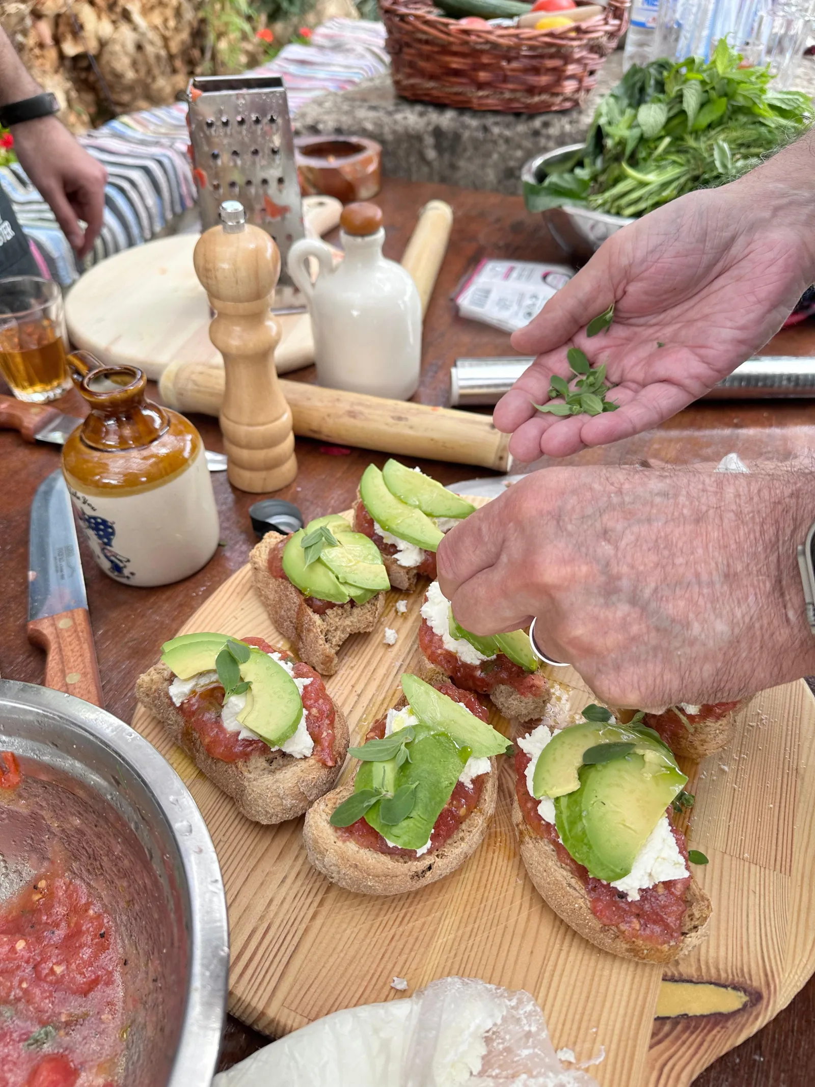Hands assembling dakos on a wooden board beside a grater, pepper mill and bowl of grated tomato