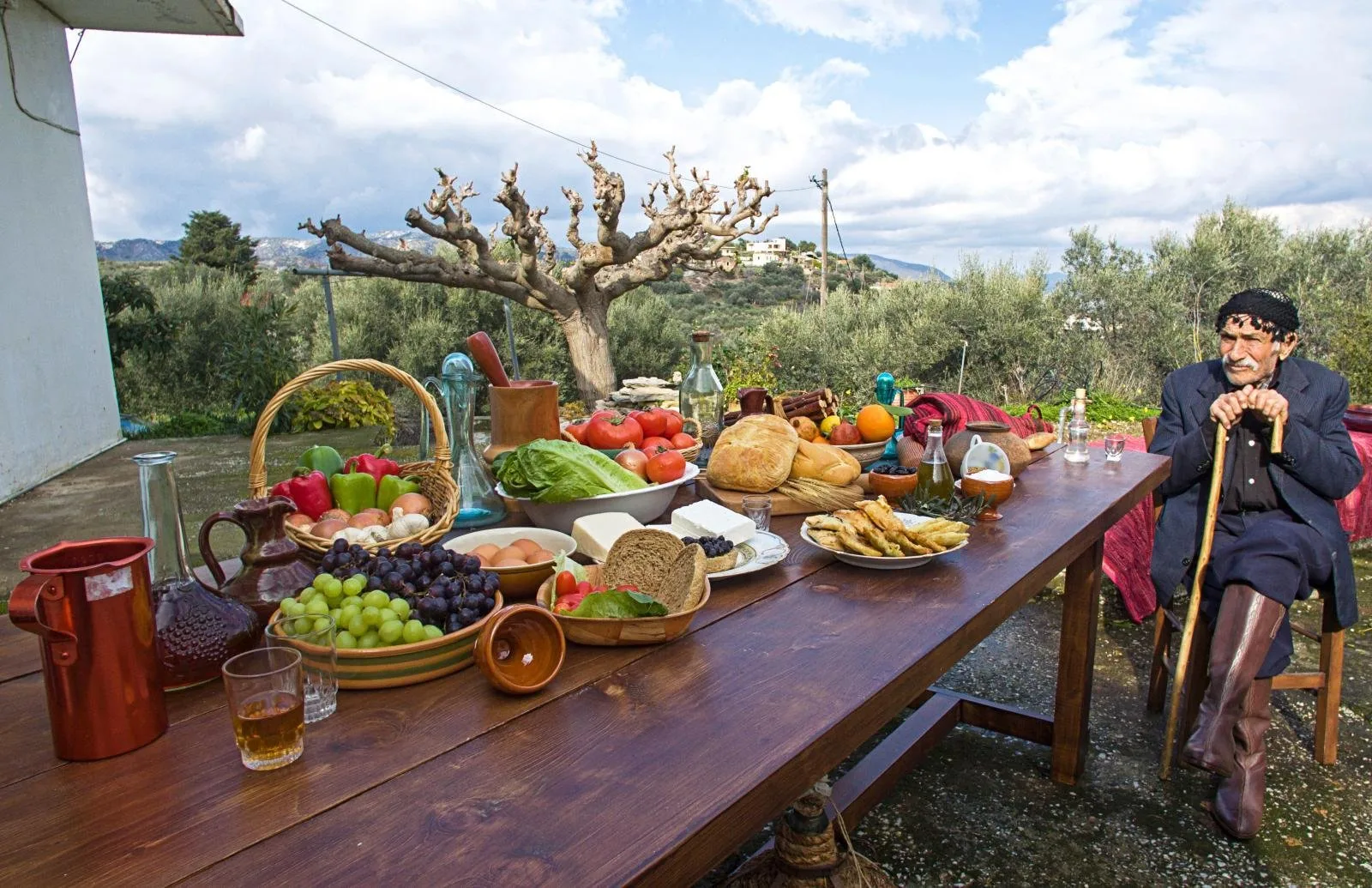 A long wooden table under an olive tree loaded with fruit, bread and salads, an elderly Cretan man seated at the far end