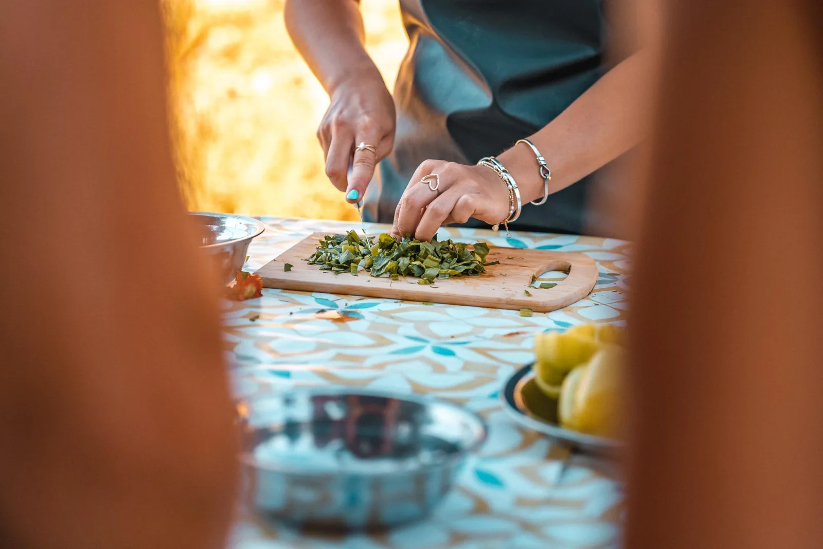 Hands chopping wild greens on a board at a tiled table, a lemon and a small pan alongside